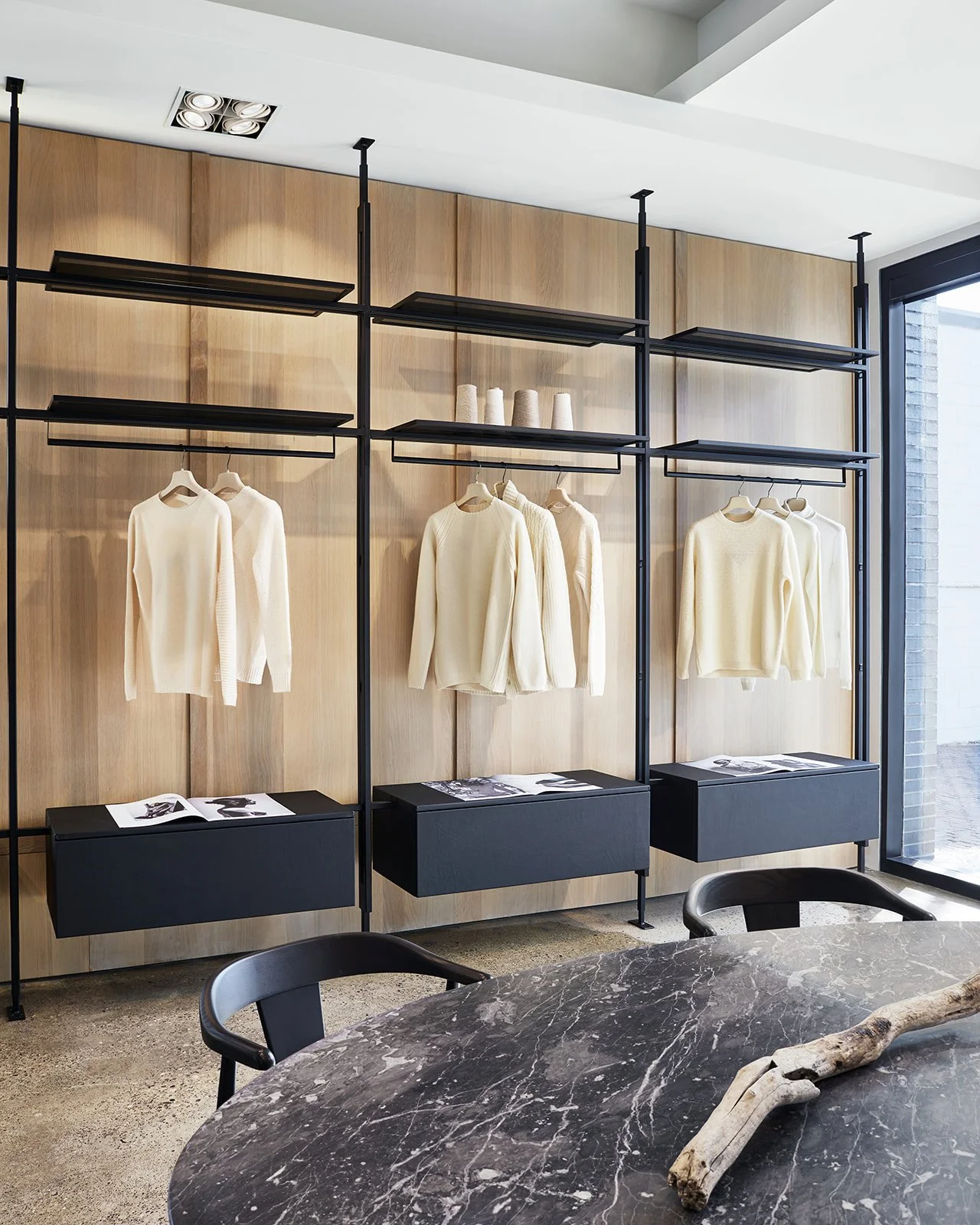 Display of white sweaters hanging on black metal racks in a modern store with a wooden wall background and a marble table in the foreground.