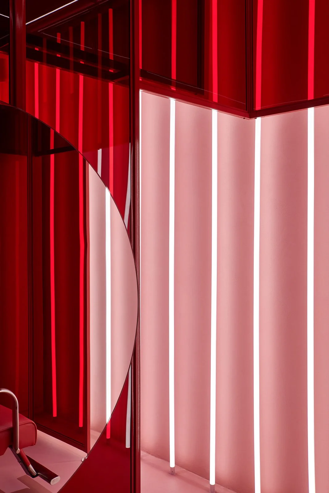 A close-up architectural detail showing a circular mirror overlapping red glass panels, reflecting the glow of vertical LED strip lights in a bold, contemporary wellness environment.