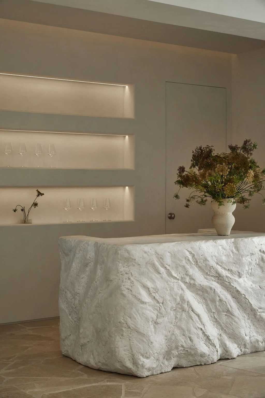 A custom sculpted plaster bar with a raw, rock-like texture in a minimalist hospitality space, featuring built-in illuminated shelving, limewash walls, and a floral arrangement.
