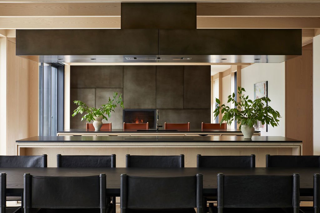 An open-concept kitchen and dining space designed for slow living, featuring a long black dining table, a light wood kitchen island beneath a dramatic dark metal range hood, and a custom fireplace in the background.
