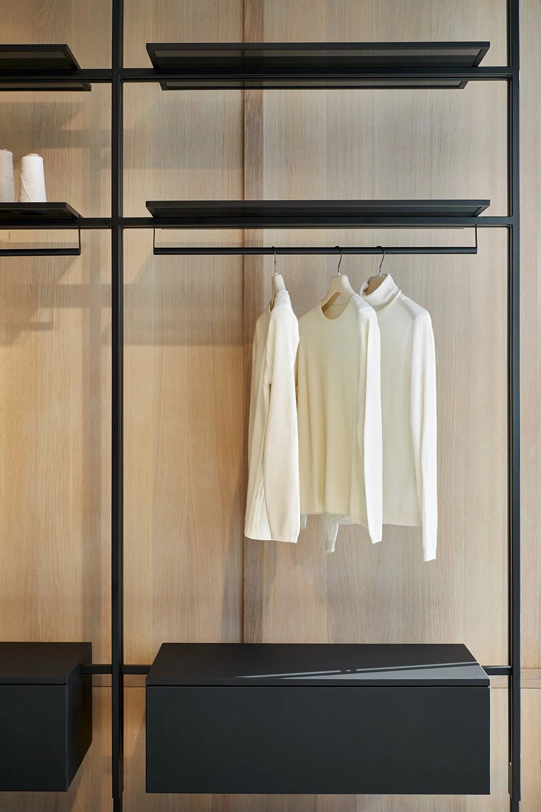 Wooden closet with black metal frame, three white sweaters hanging on a black rod, and black shelves on either side.