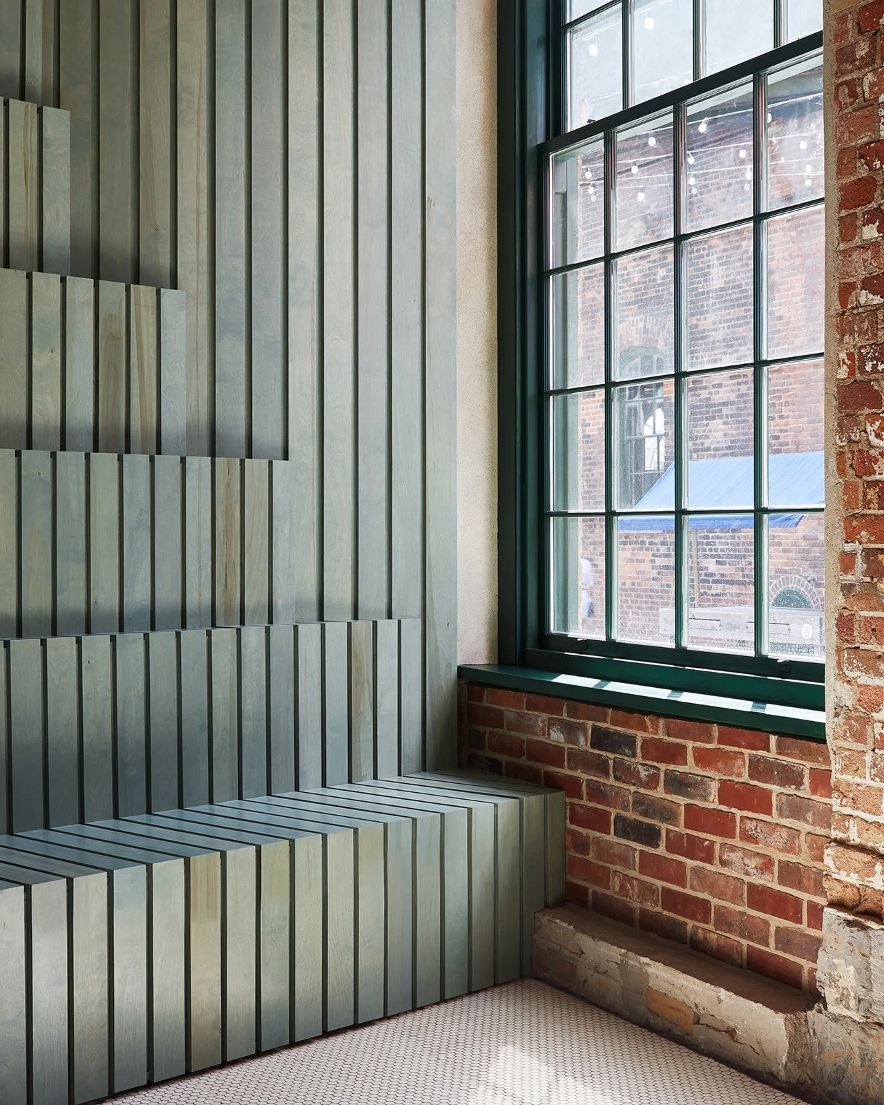 Interior corner with a large window, exposed brick wall, and a custom greenish wood-panel seating area.