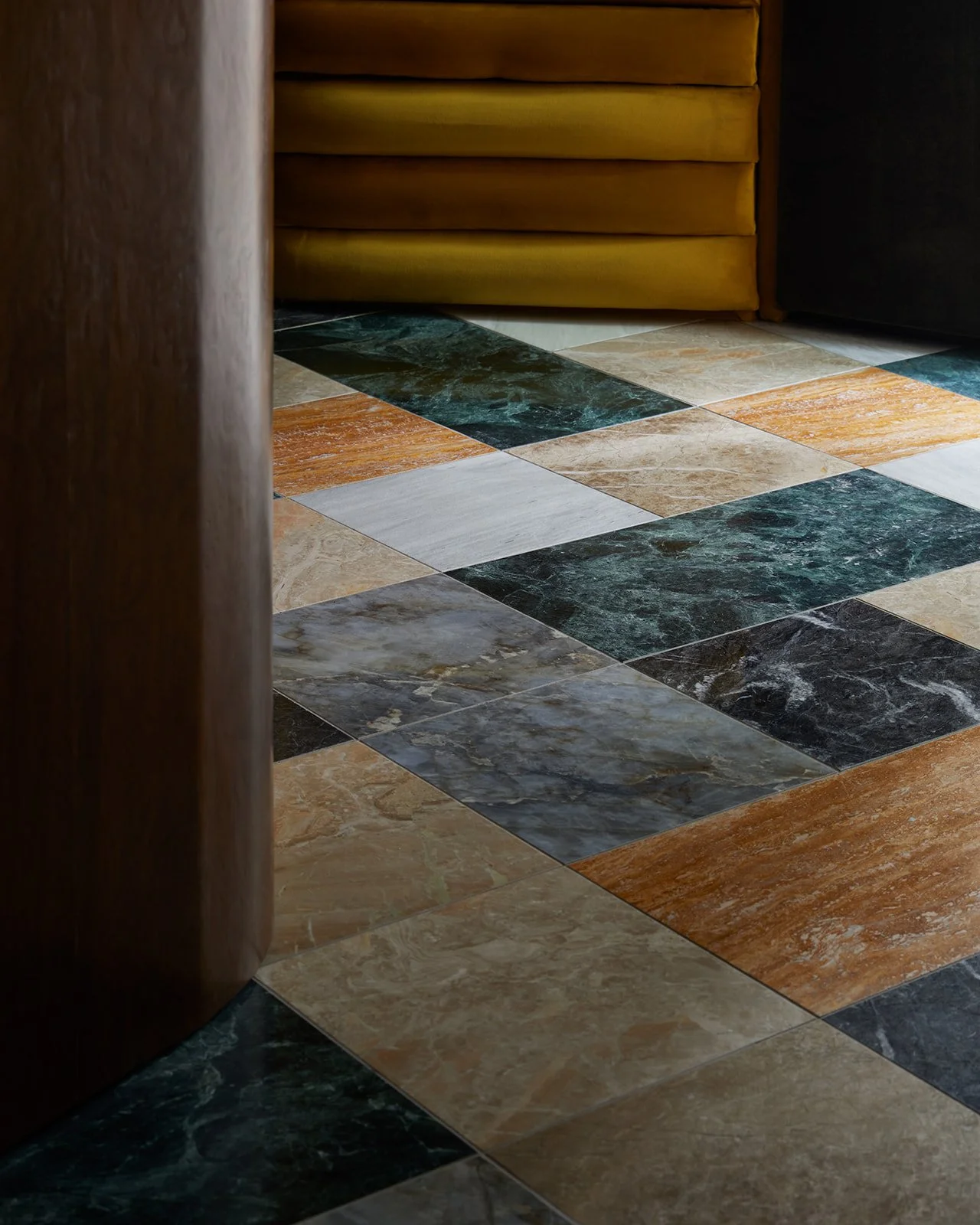 An editorial close-up of luxury multi-colored marble checkerboard flooring in deep greens, tans, and greys, contrasting with the base of a curved wood desk and gold velvet seating.