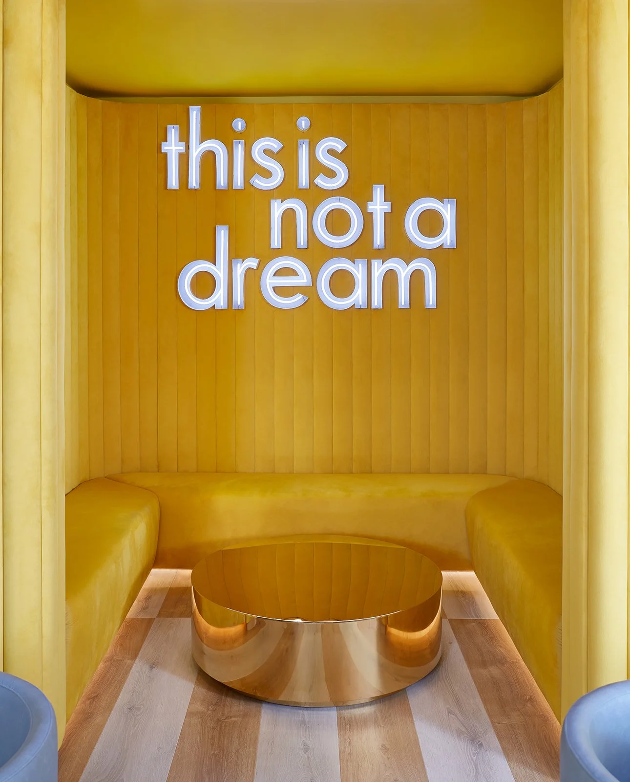 Bright yellow interior with a neon sign that reads "this is not a dream" hanging on the wall. There is a yellow bench curved around a round, reflective, golden table.