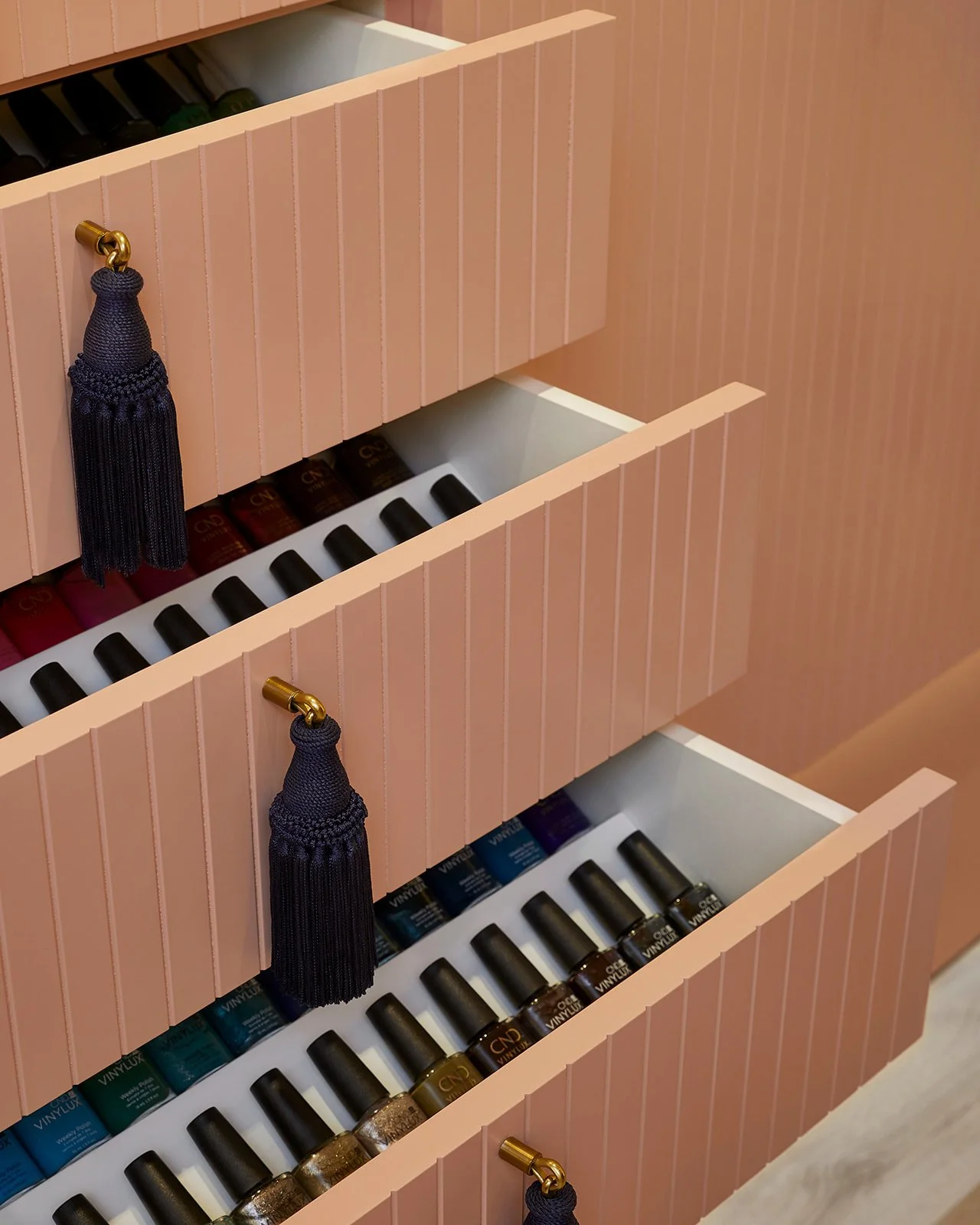 Pink nail polish storage cabinet with black tassel hooks and various bottles of nail polish inside.