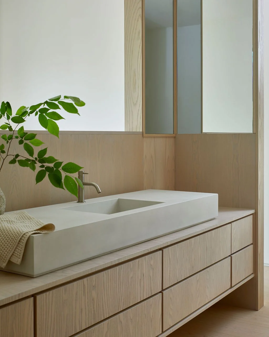 A modern, minimalist bathroom design featuring a long, custom concrete sink basin resting on a floating light wood vanity with seamless drawers, surrounded by wood-paneled walls and corner mirrors.