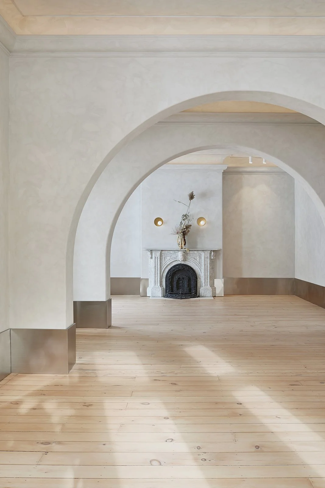 A minimalist interior room with arched doorways, light-colored walls, wooden flooring, and a white fireplace decorated with a vase of dried flowers. Small wall sconces are mounted above the fireplace.