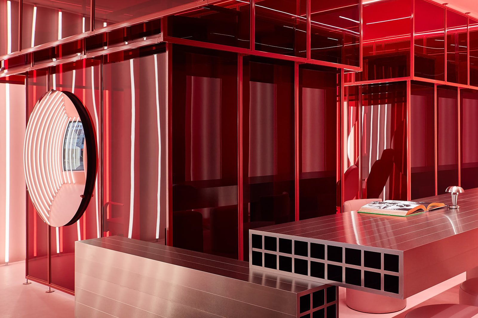 A wide view of the hospitality-inspired reception and lounge area at Pout X, featuring a monumental floating metallic desk, pink cylindrical stools, and immersive red glass architectural partitions.