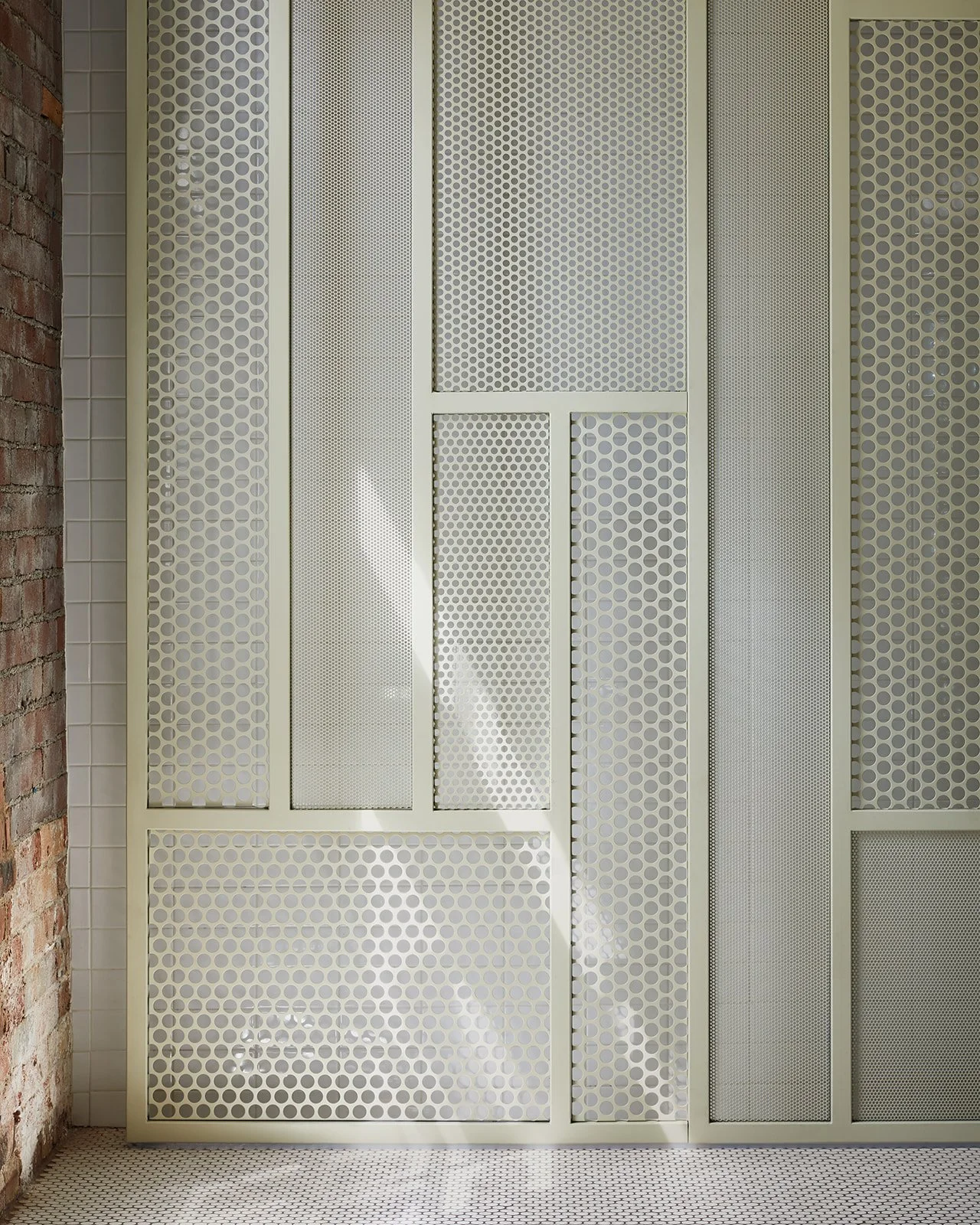 Interior decorative wall with perforated metal panels in various sizes, adjacent to an exposed brick wall, with a small tiled floor.