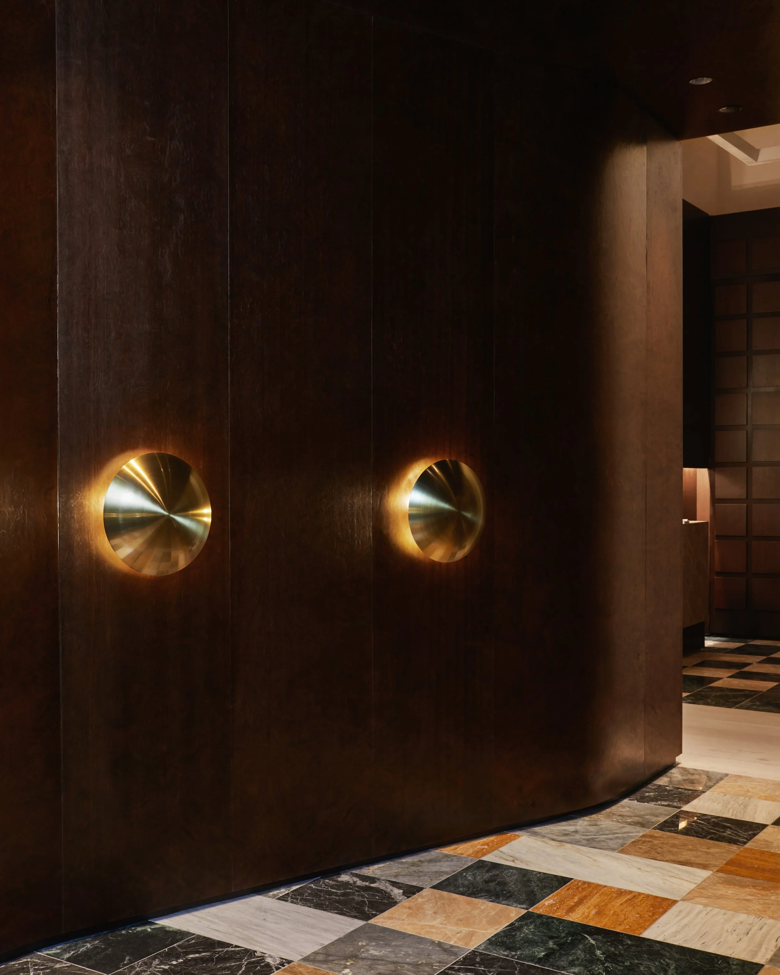 A sweeping curved corridor finished in rich dark wood paneling, illuminated by modern brass dome wall sconces, leading over custom multi-colored marble checkerboard floors.