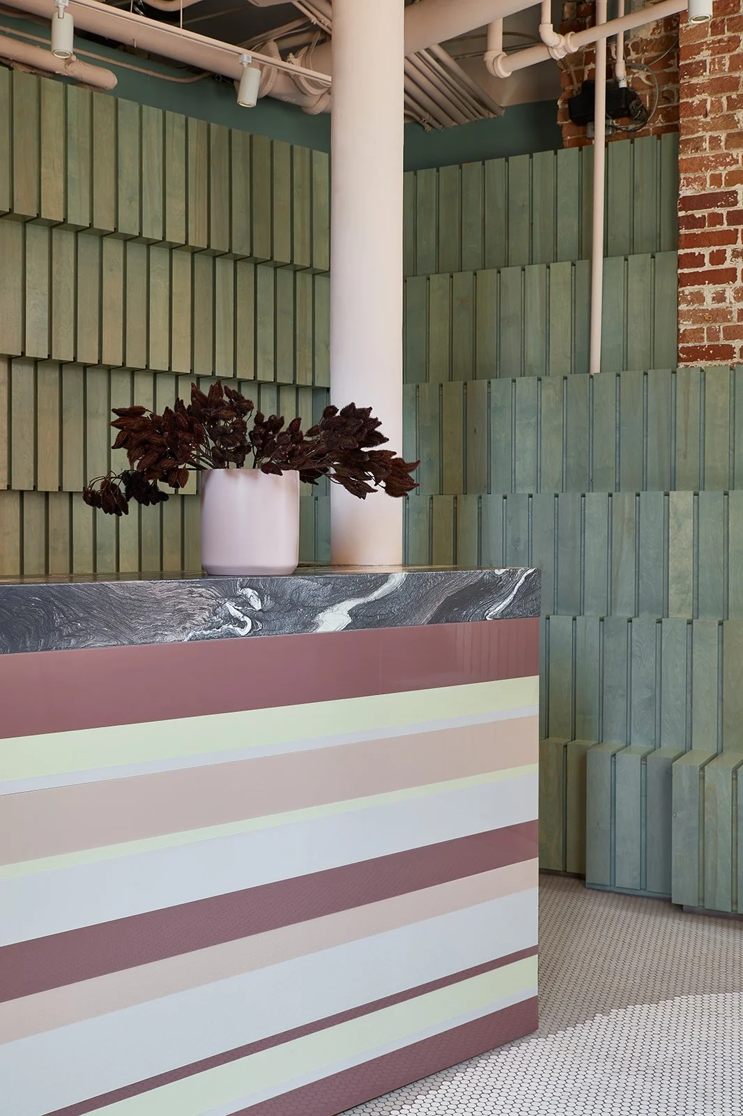 A modern interior with a striped pastel-colored counter, a white vase with dark red leaves, a green wood-paneled wall, and an exposed brick wall in the background.
