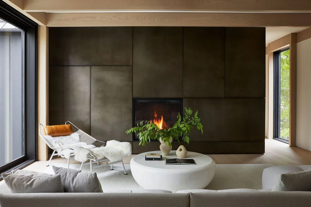 A serene living area in a private retreat featuring a monumental dark metal paneled fireplace wall, a sculptural white coffee table, and a relaxing lounge chair styled with a sheepskin throw, framed by light wood ceiling beams.