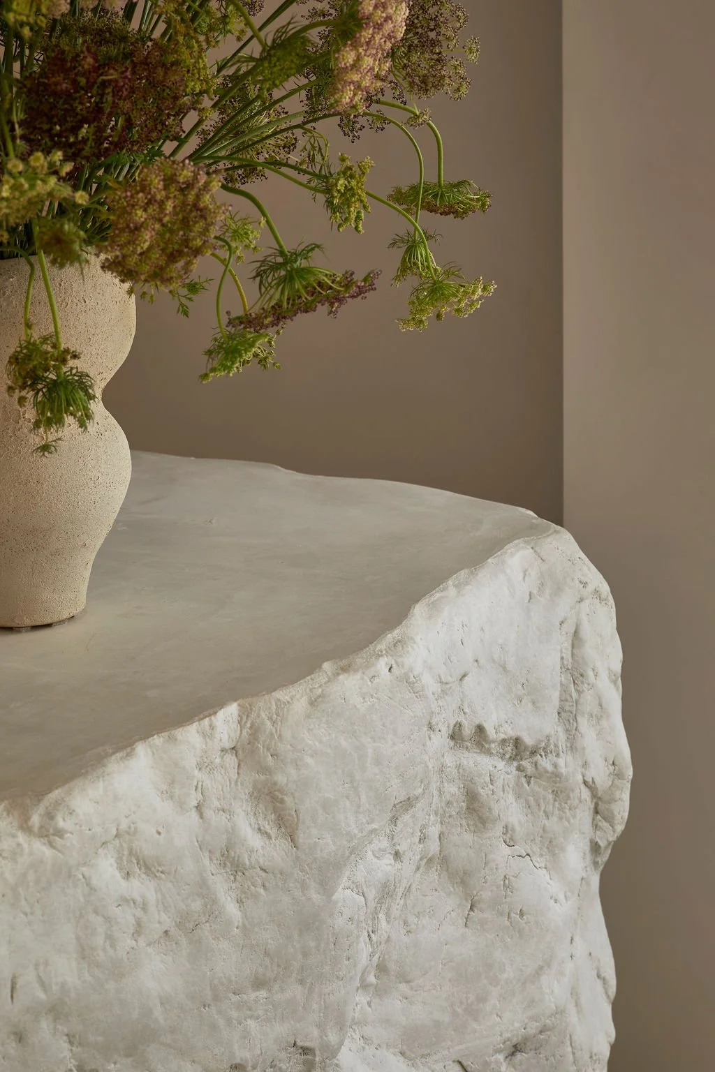A custom sculpted plaster bar with a raw, rock-like texture in a minimalist hospitality space, featuring built-in illuminated shelving, limewash walls, and a floral arrangement.