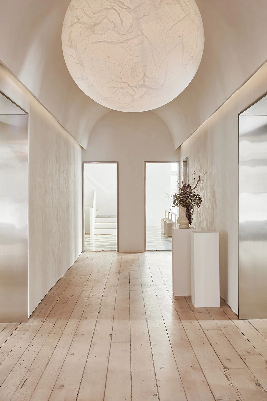 Minimalist hallway with light wooden floor, white walls, metal storefronts, a large round ceiling light, and a white pedestal with a floral arrangement.