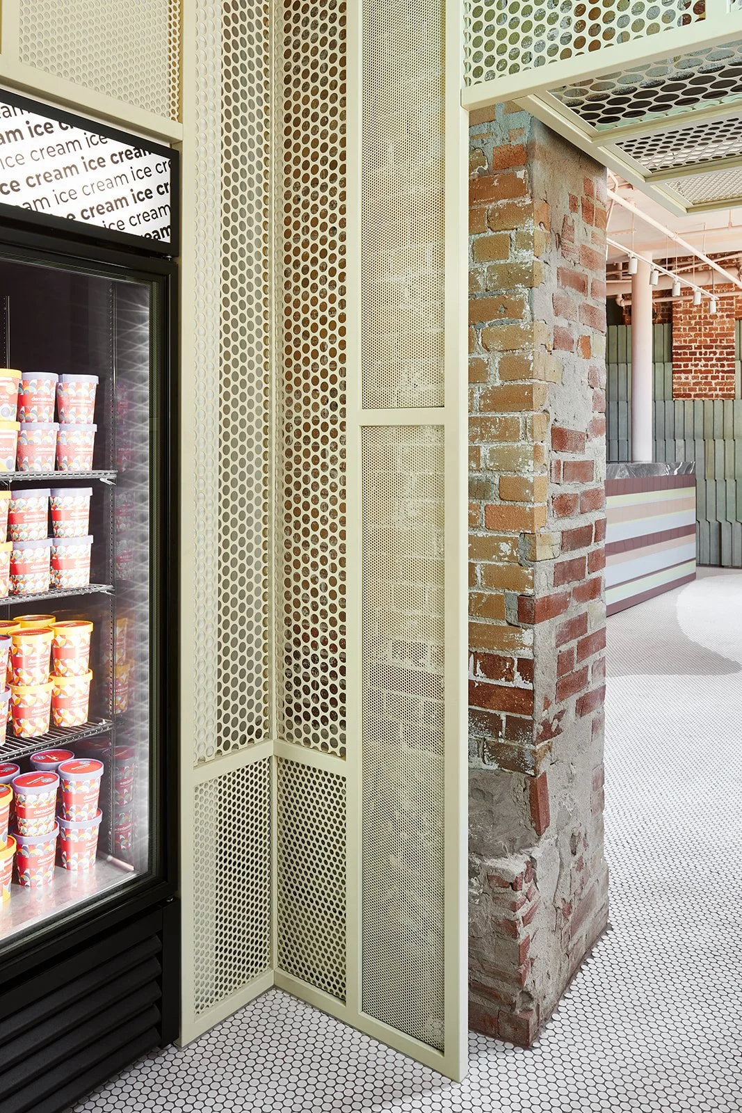 Ice cream freezer with cups next to a cream-colored perforated metal divider, brick wall, and patterned white tile flooring.