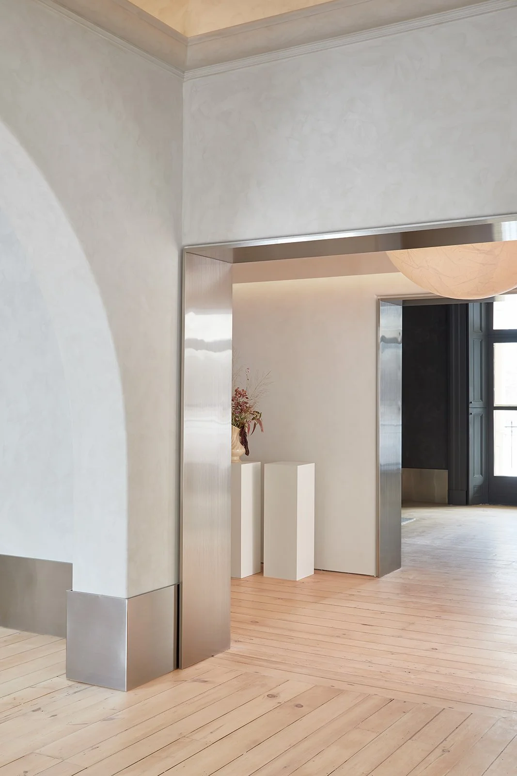 Minimalist interior of a room with light-colored wooden floors, white walls, a wall-mounted light fixture, and a black window frame with multiple panes.