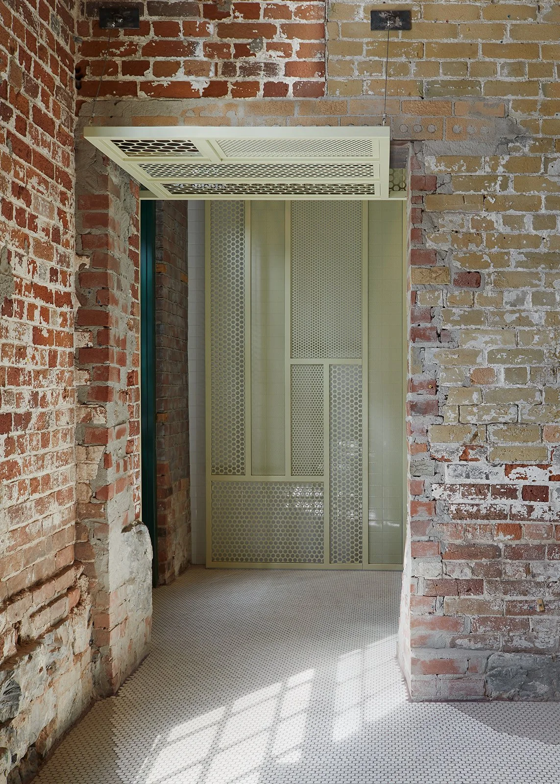 An interior space with exposed brick walls, a white patterned door, and sunlight casting shadows on a small tiled floor.