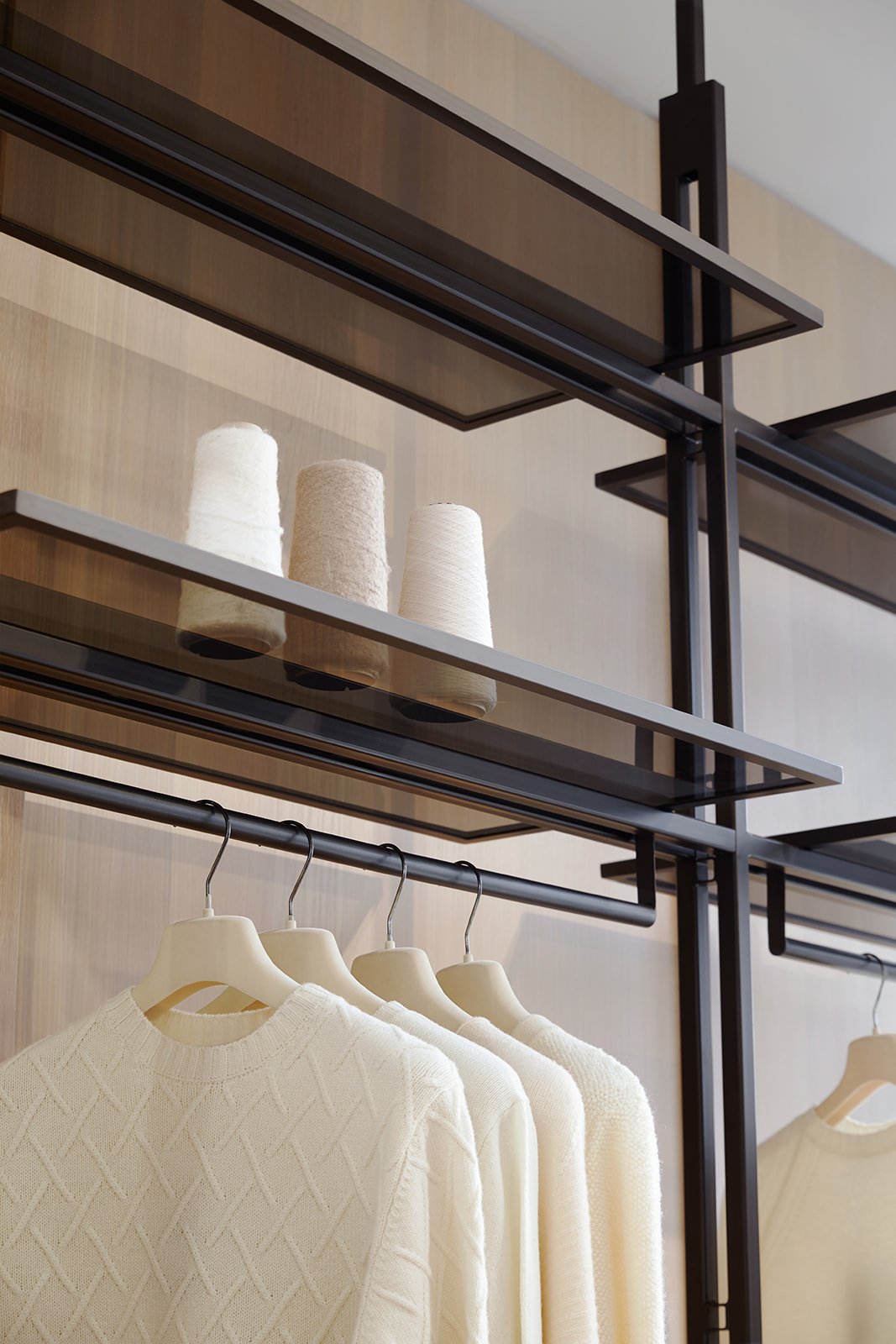 Cream-colored sweaters hanging on a black clothing rack in a retail store with wooden and metal shelving above, containing beige and white yarn spools.