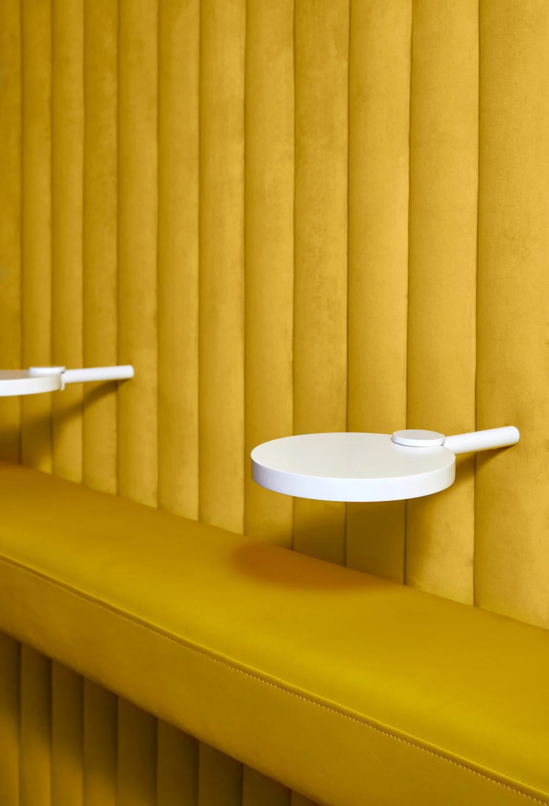 Yellow upholstered wall with white circular side tables attached, two on the right side of the image.