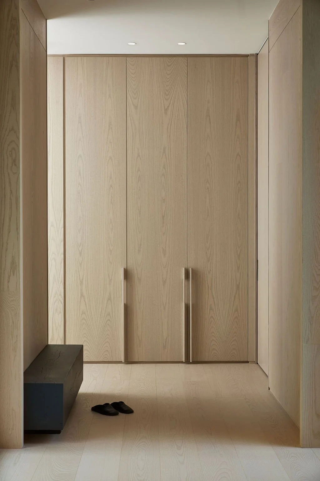 A serene, minimalist entryway in a private retreat featuring floor-to-ceiling seamless wood closets with custom integrated handles, light wood flooring, and a contrasting dark structural bench.