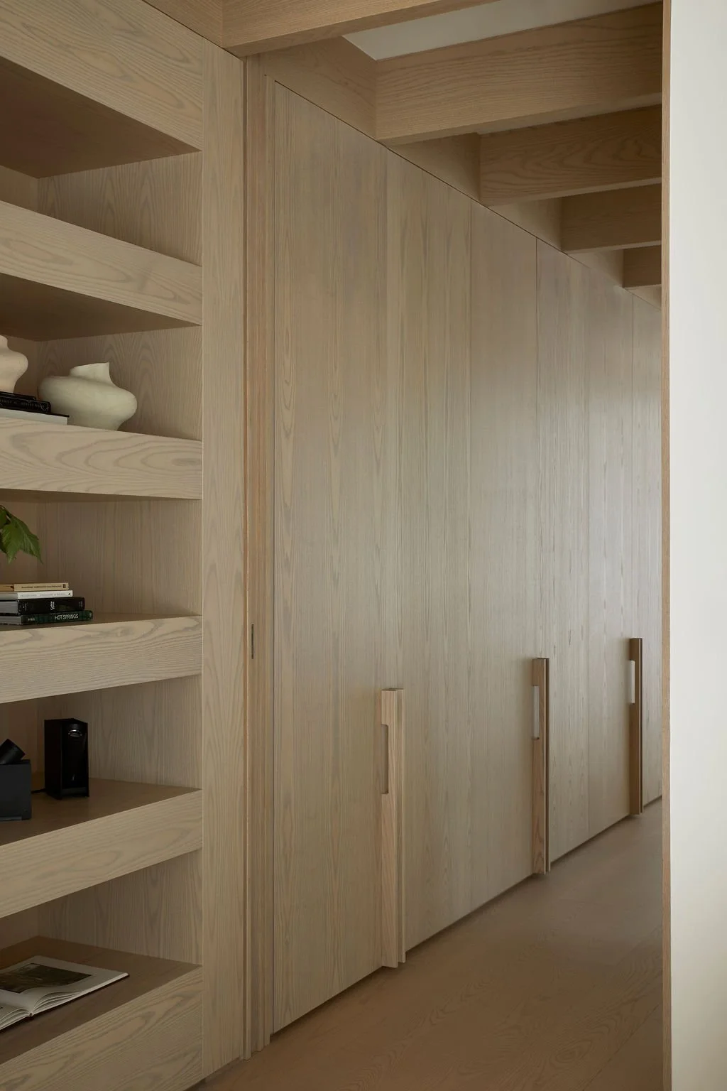 A serene hallway in a bespoke retreat showcasing extensive custom light wood millwork, including seamless closets with integrated wooden handles, built-in display shelving, and exposed ceiling beams.