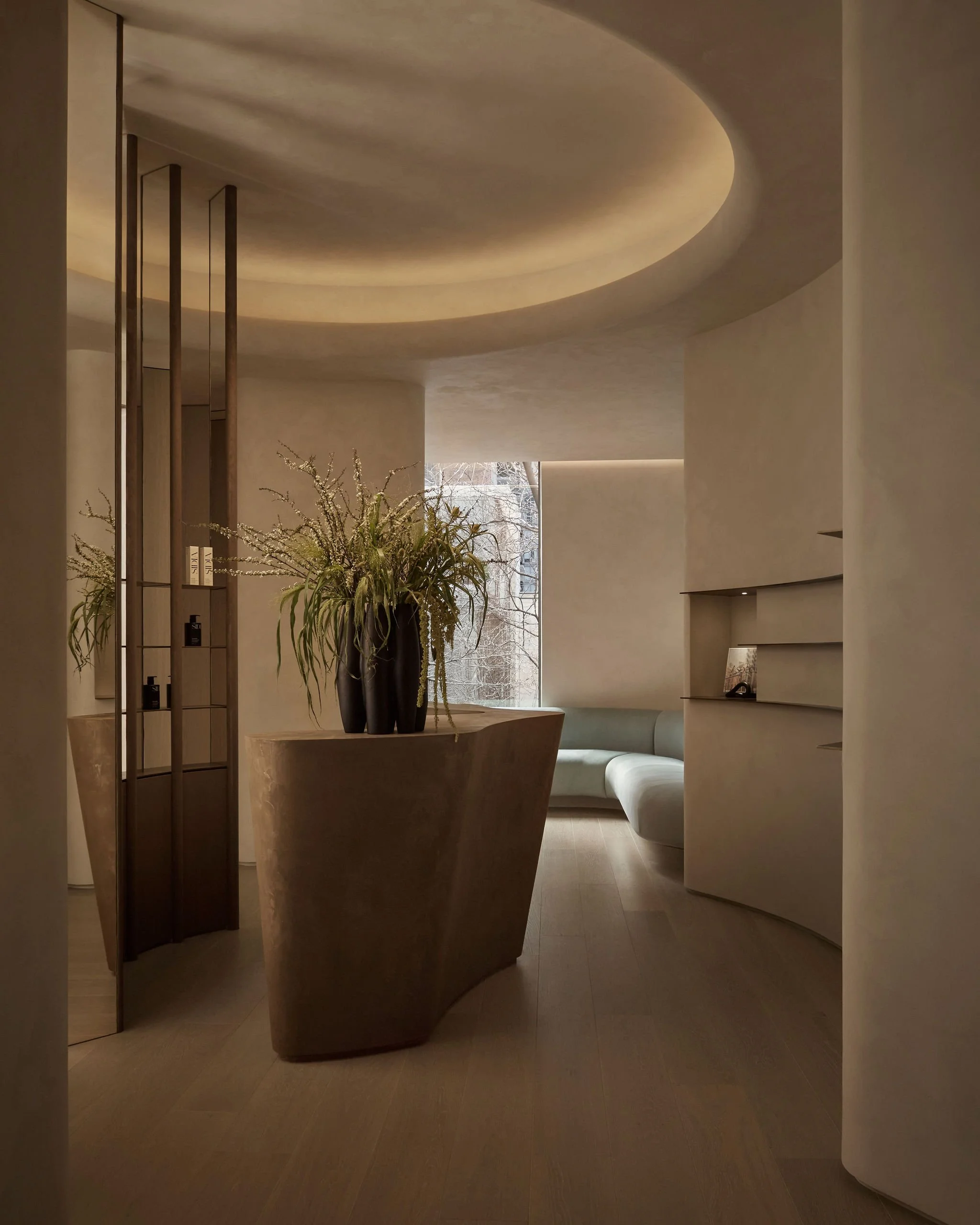 Modern minimalist clinic reception area featuring a sculptural curved desk, warm limewash plaster walls, a mirrored wood room divider with retail displays, and a recessed ceiling with ambient lighting
