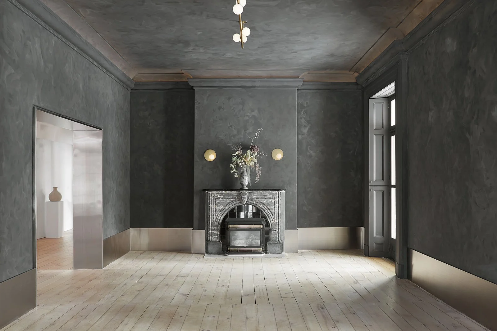 A minimalist room with dark gray walls, a gray marble fireplace with a silver object inside, a vase with flowers on top of the fireplace, and two wall-mounted round lights on either side. There are large windows on the right side and a doorway on the left leading to another room with a white sculpture on a pedestal. The room has light-colored wooden floors and a modern ceiling light fixture.