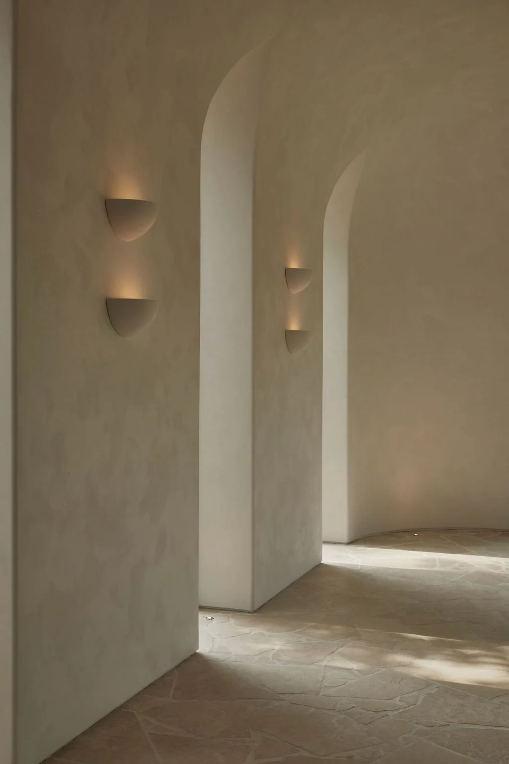 A curved interior event hall showcasing repeating arched windows, natural stone floors, and minimalist half-dome wall sconces casting warm ambient light on textured walls.