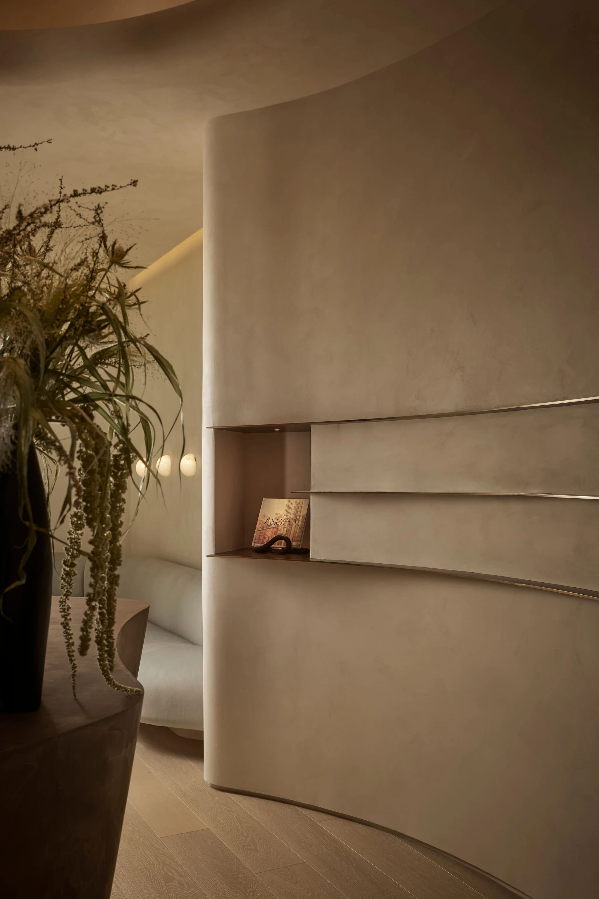 View of a thick curved limewash wall with a custom recessed display shelf, leading into a serene waiting area featuring a minimalist curved sofa and glowing wall sconces