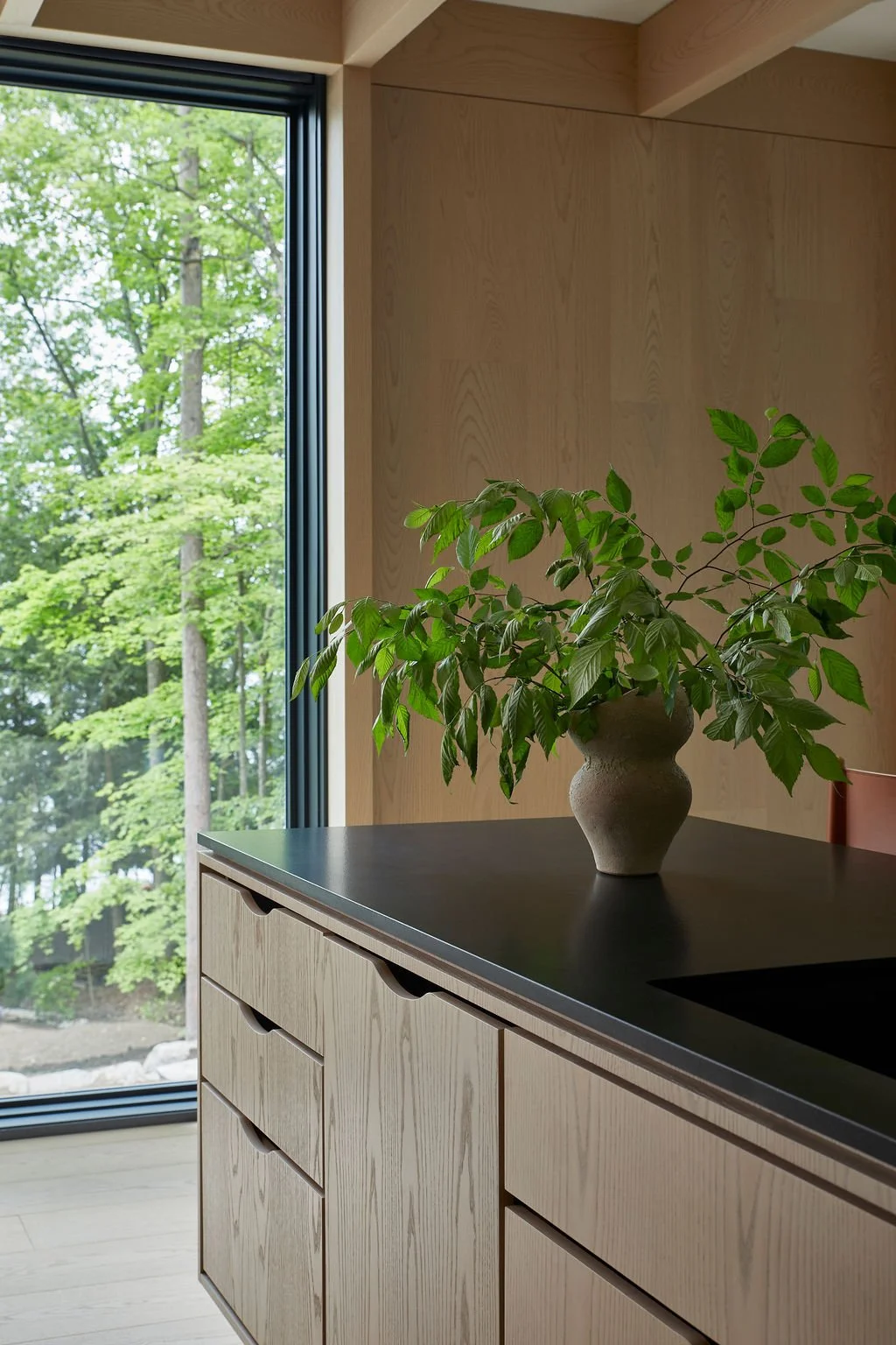 A close-up of bespoke light wood kitchen cabinetry with seamless integrated cut-out drawer pulls and a dark countertop, styled with a rustic vase and green foliage against a large window framing a dense forest.