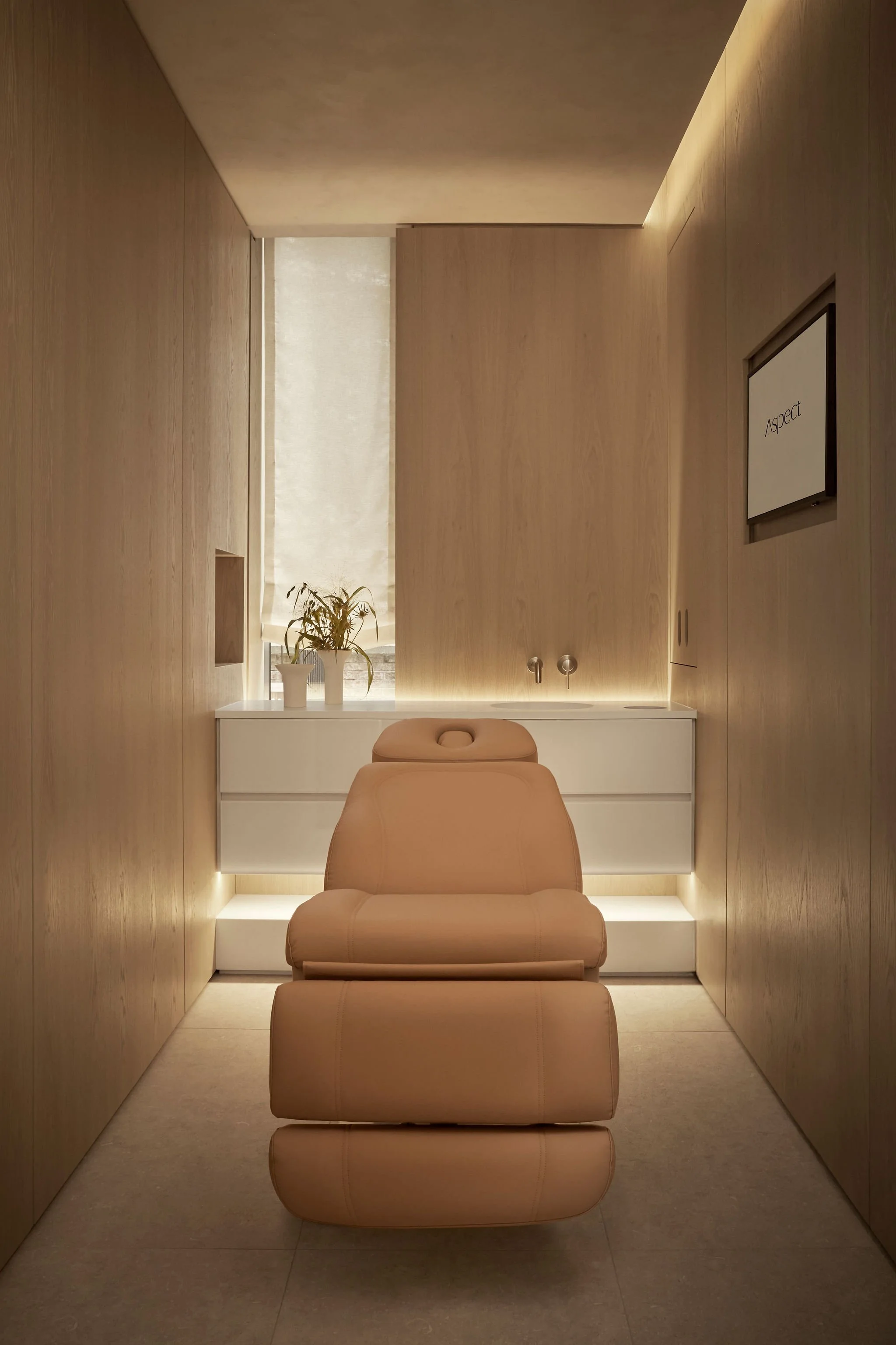 Serene surgeon's treatment room featuring warm floor-to-ceiling wood paneling, a tan leather treatment chair, a minimalist white floating vanity with modern fixtures, and soft ambient under-cabinet lighting