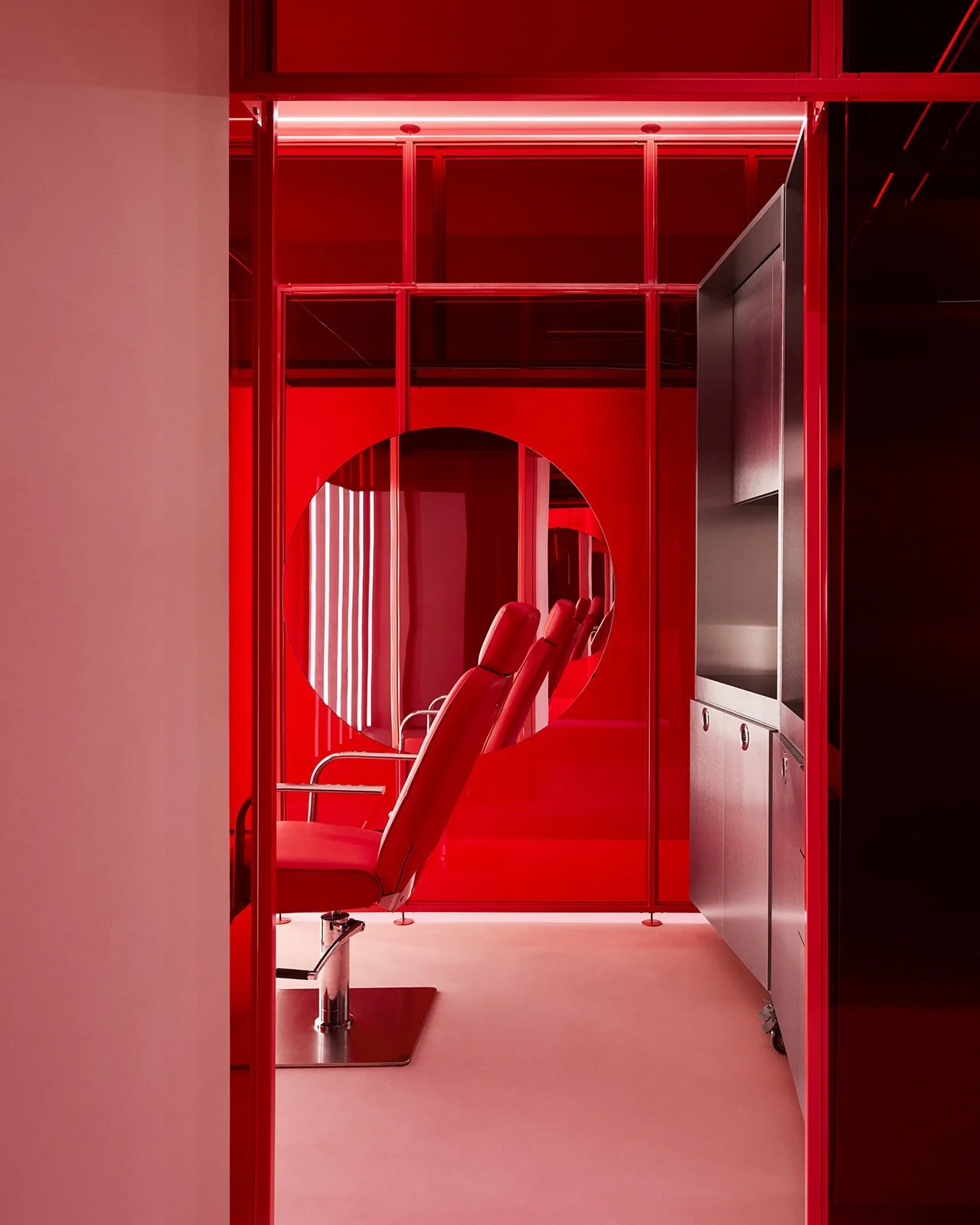 A bold, anti-clinical treatment room at Pout X medical spa featuring translucent red glass walls, a circular mirror, a red reclining treatment chair, and sleek stainless steel clinical cabinetry.