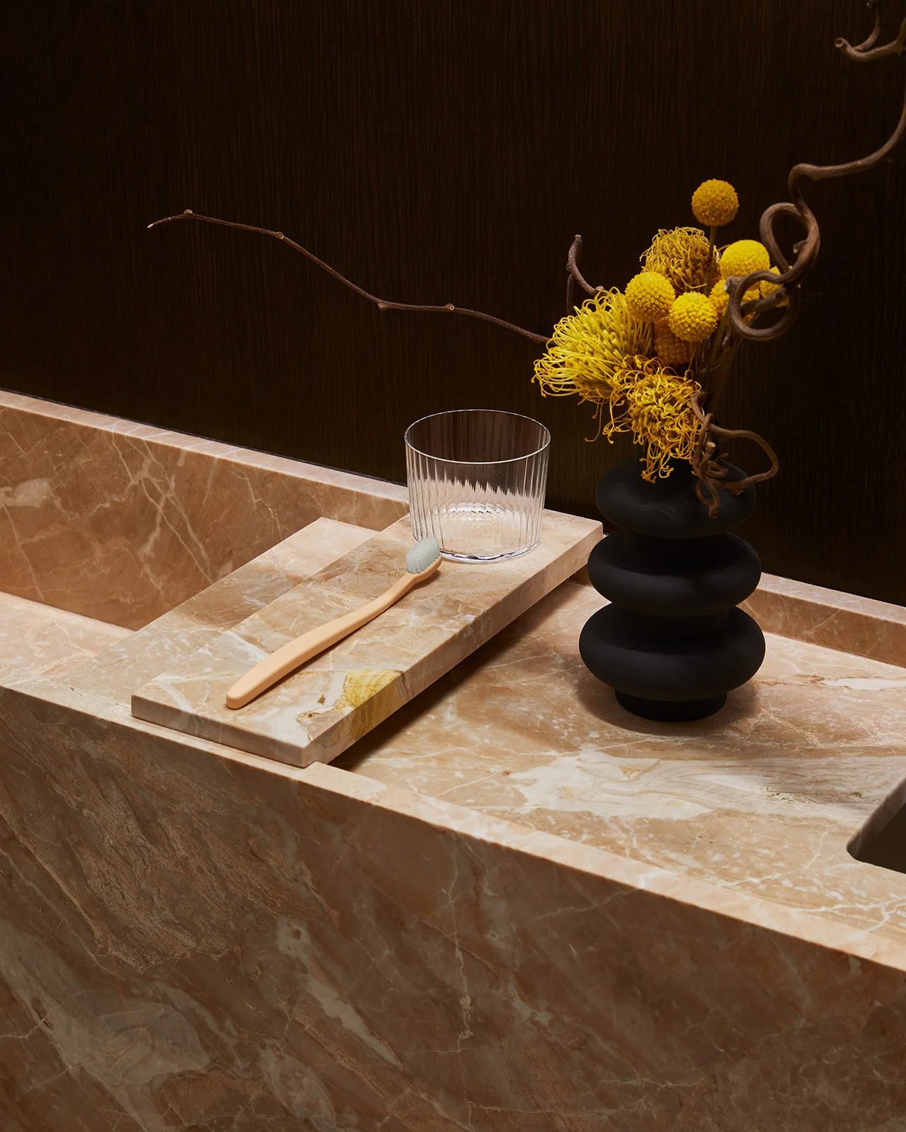 A close-up detail of a bespoke marble sink in a luxury dental clinic, styled with a ribbed glass cup, a bamboo toothbrush, and a sculptural black vase.