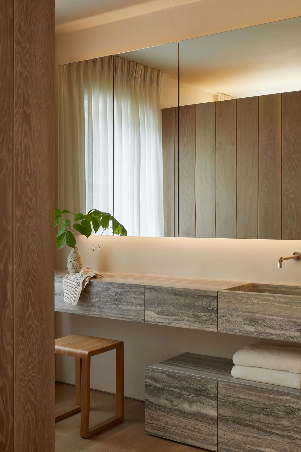 A luxury bathroom vanity carved from layered travertine stone, featuring a dedicated makeup seating area with a curved wooden stool, under-cabinet lighting, and seamless mirrored upper cabinets.