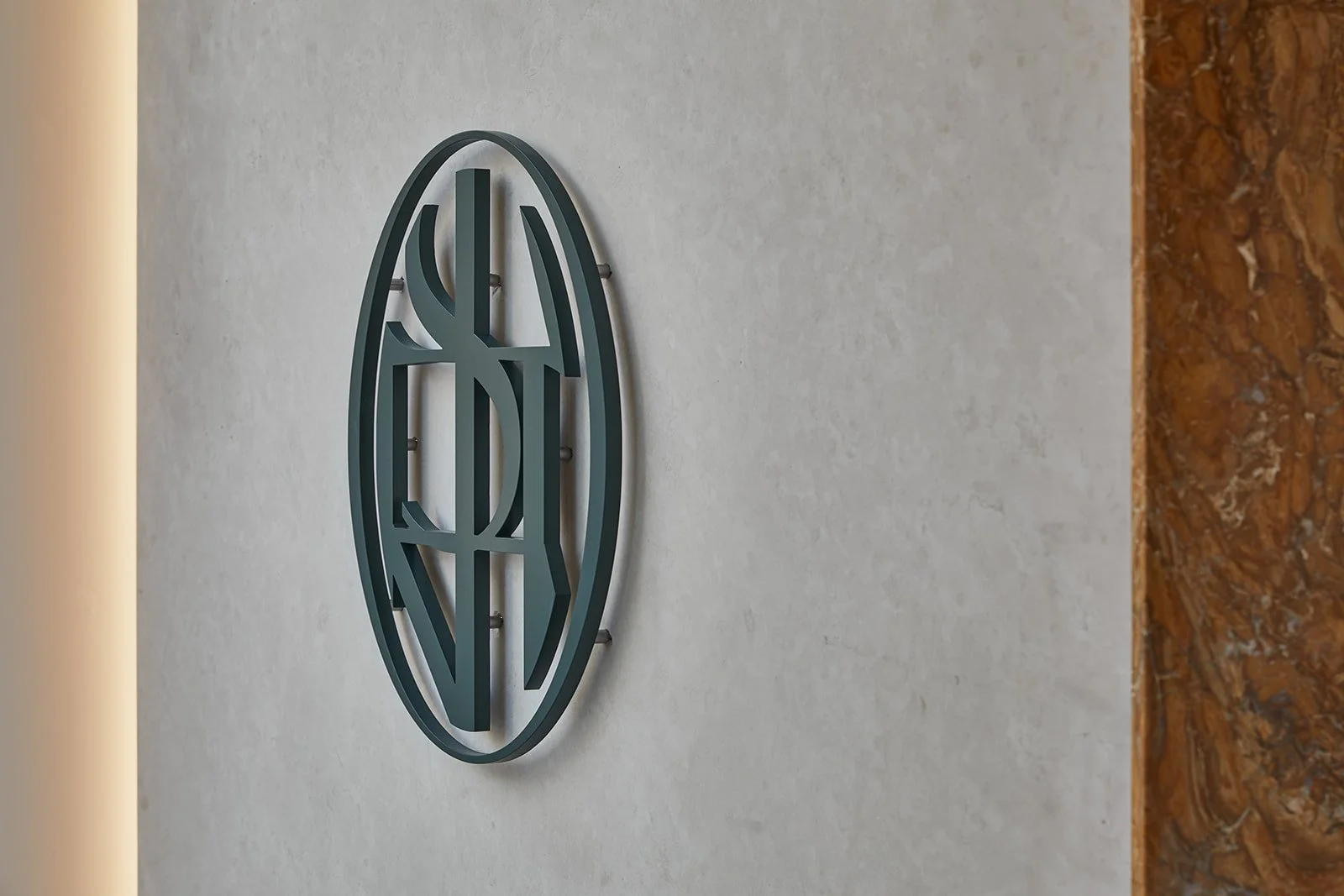 A close-up architectural detail of the custom dark metal monogram signage at Paste boutique dental clinic, mounted with standoffs on a textured light stone wall and accented by warm ambient lighting and rich brown marble.