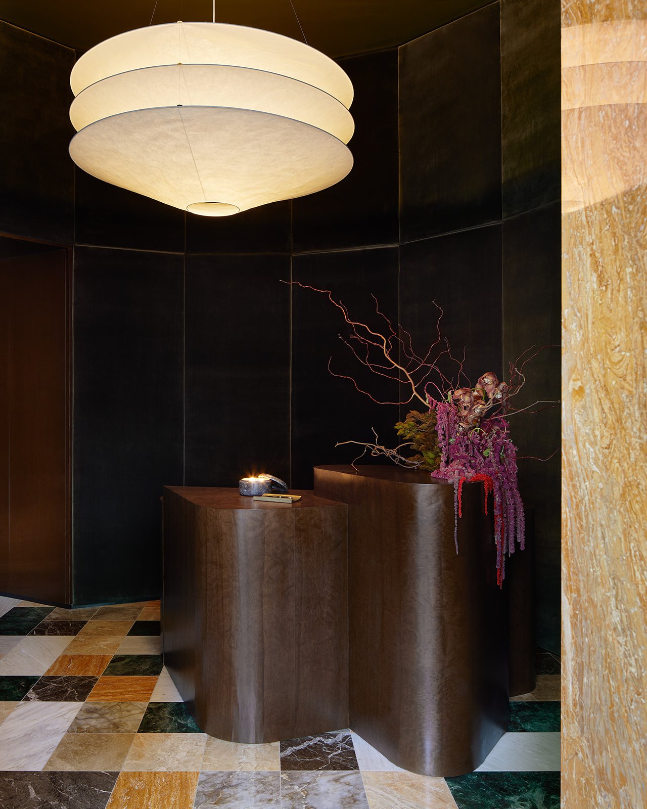 A hospitality-inspired reception area at Paste boutique dental clinic, featuring a custom, staggered burl wood desk, a large paper lantern pendant light, and multi-colored marble checkerboard flooring against dark textured walls.