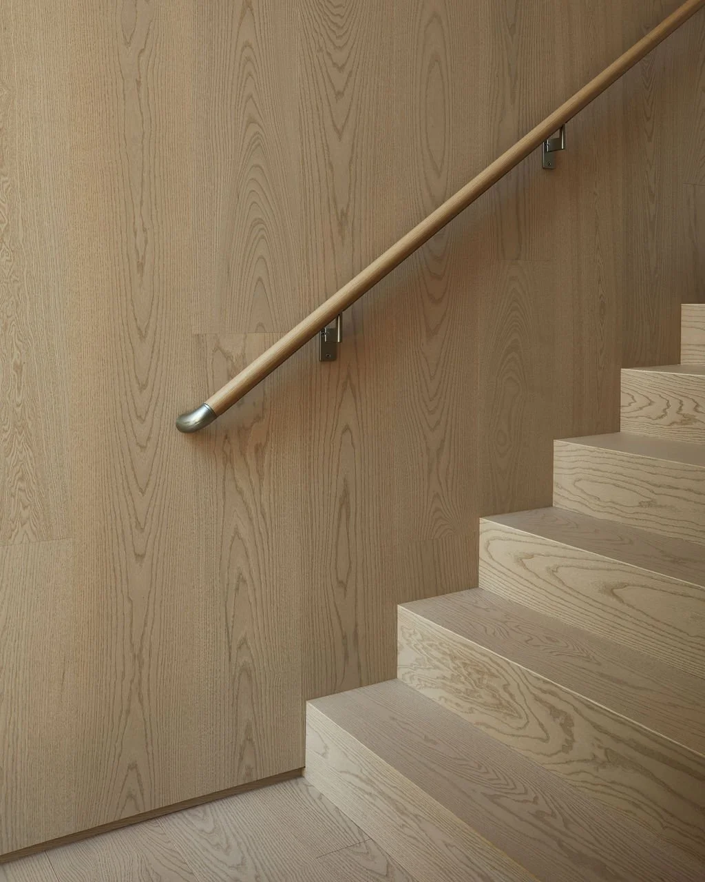A minimalist light wood staircase blending seamlessly with matching floor-to-ceiling wood wall paneling in a private recreational residence, accented by a simple wooden handrail.
