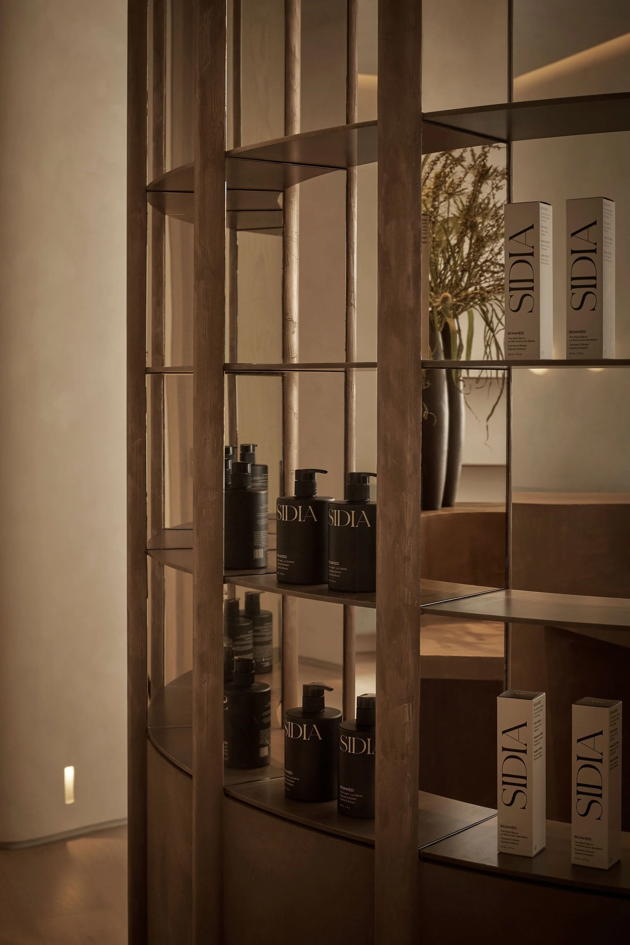 Close up of a custom floor-to-ceiling mirrored bronze plaster shelving unit displaying boutique skincare products in a modern wellness clinic reception area
