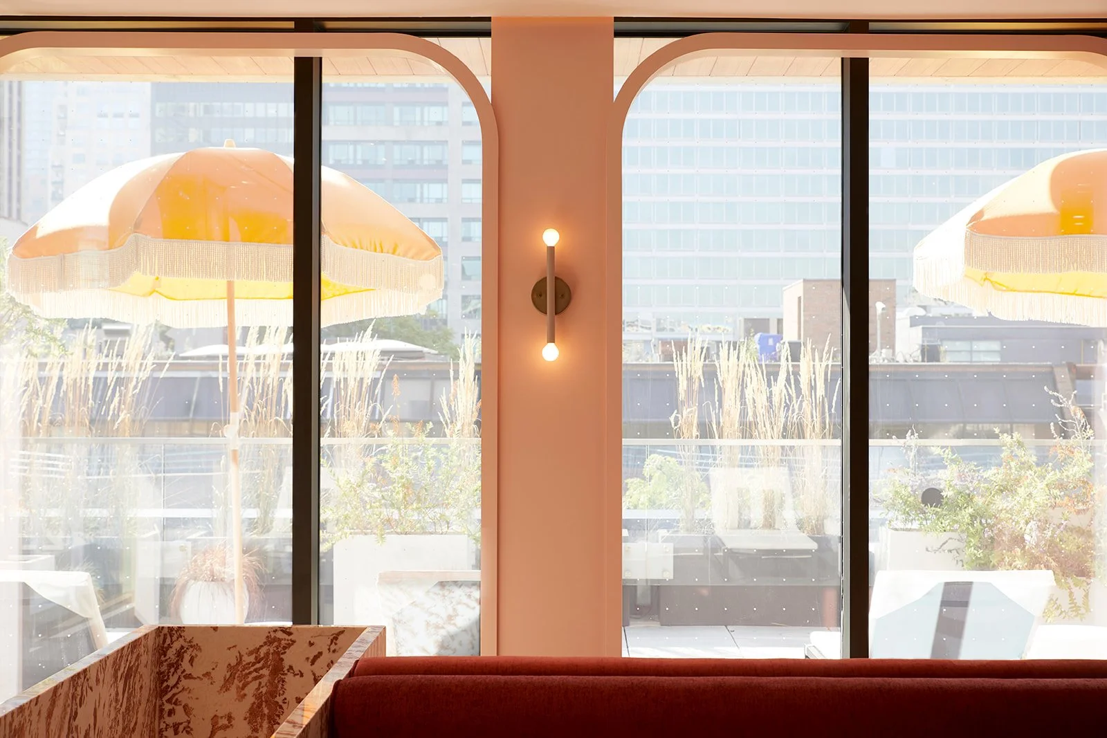 Interior view of a cafe with a large window, yellow patio umbrellas outside, and city buildings in the background. Soft lighting inside with a wall-mounted light fixture. Red seating in the foreground.