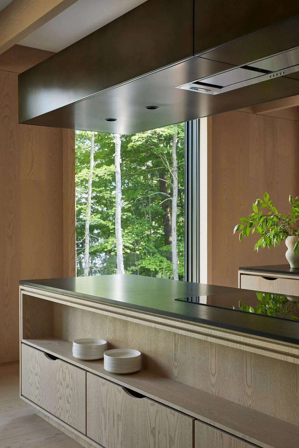 A nature-inspired kitchen in the Souno recreational residence showcasing a custom light wood island with open shelving and a dark countertop, positioned beneath a sleek metal range hood and a large window framing a forest view.