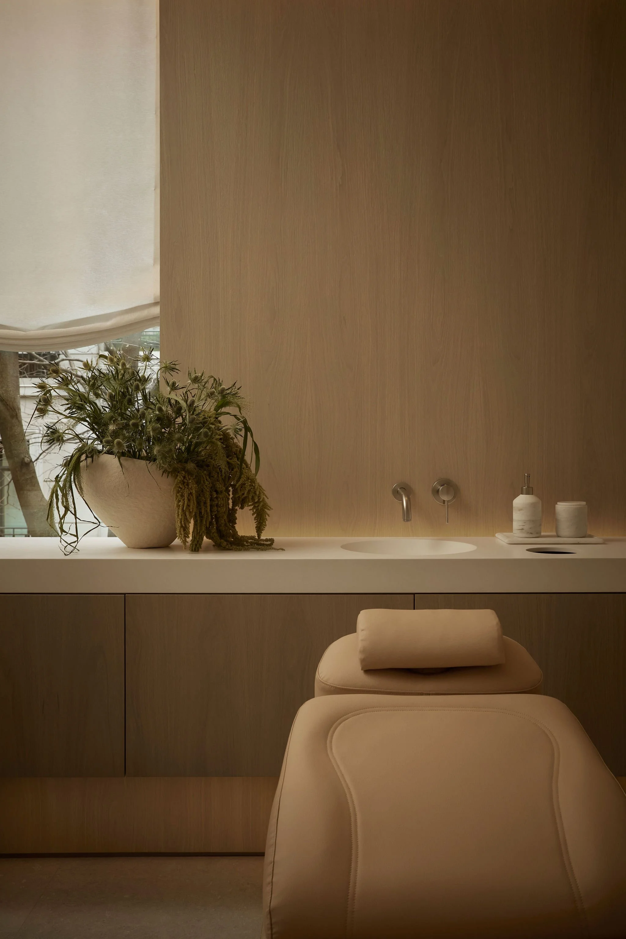 Serene boutique clinic treatment room featuring a tan leather facial bed, a sleek white sink counter with wall-mounted fixtures, and a large textured planter with cascading greenery set against warm wood paneling
