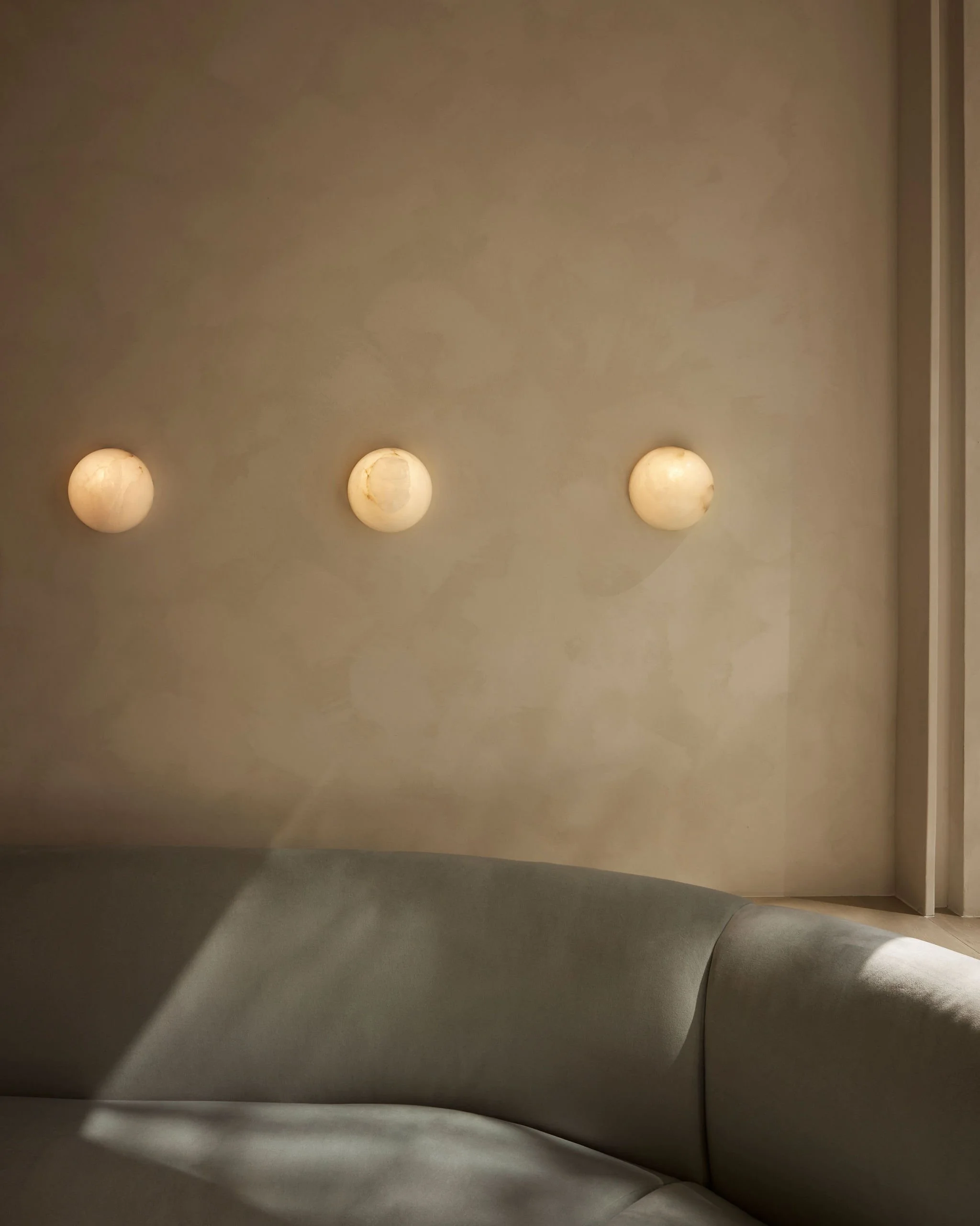 Serene minimalist waiting room interior featuring a curved grey-green sofa set against a textured neutral wall, illuminated by three round glowing alabaster wall sconces