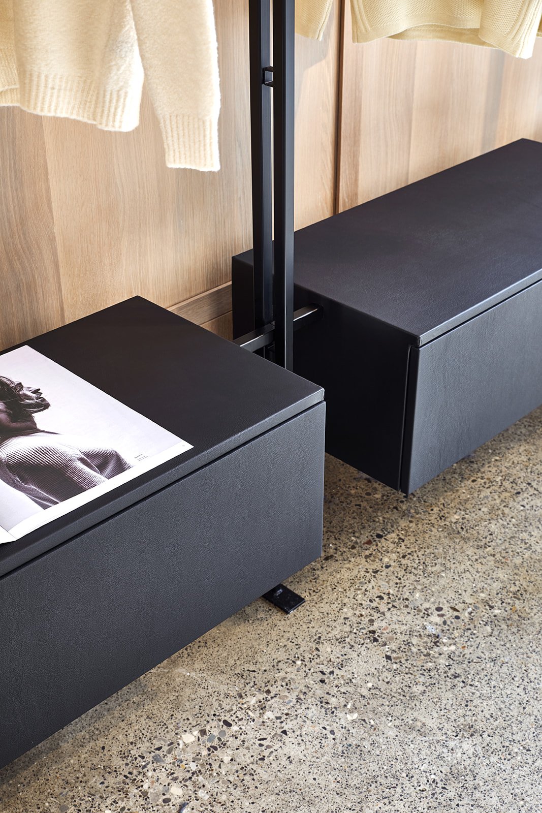 Black bench with a magazine on top, next to another black bench, in front of wooden wall panels and beige curtains, with a speckled concrete floor.