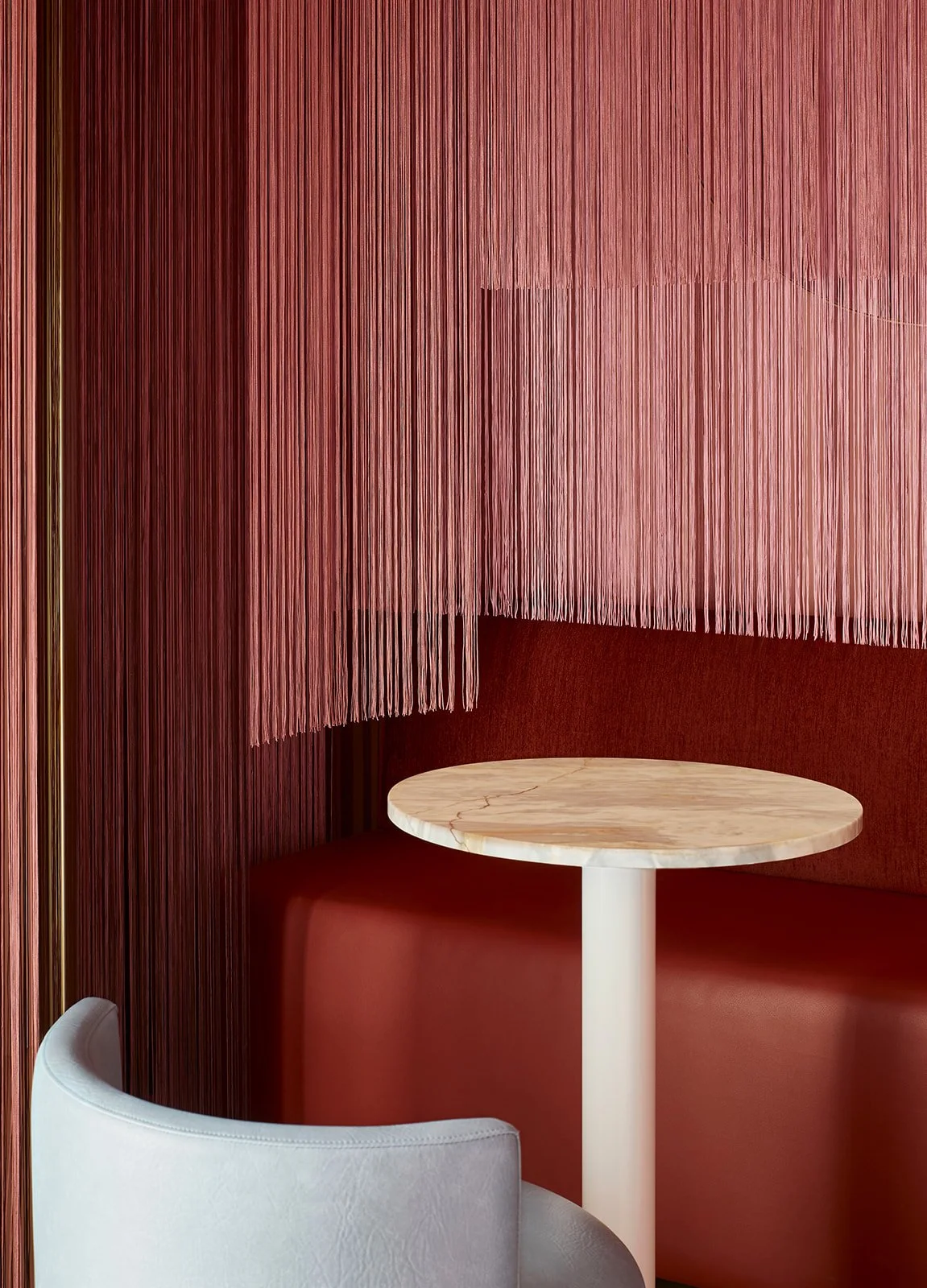 A cozy corner of a restaurant or cafe with a round marble table, a cushioned red booth, a light gray chair, and a backdrop of pink vertical string curtains and textured red walls.