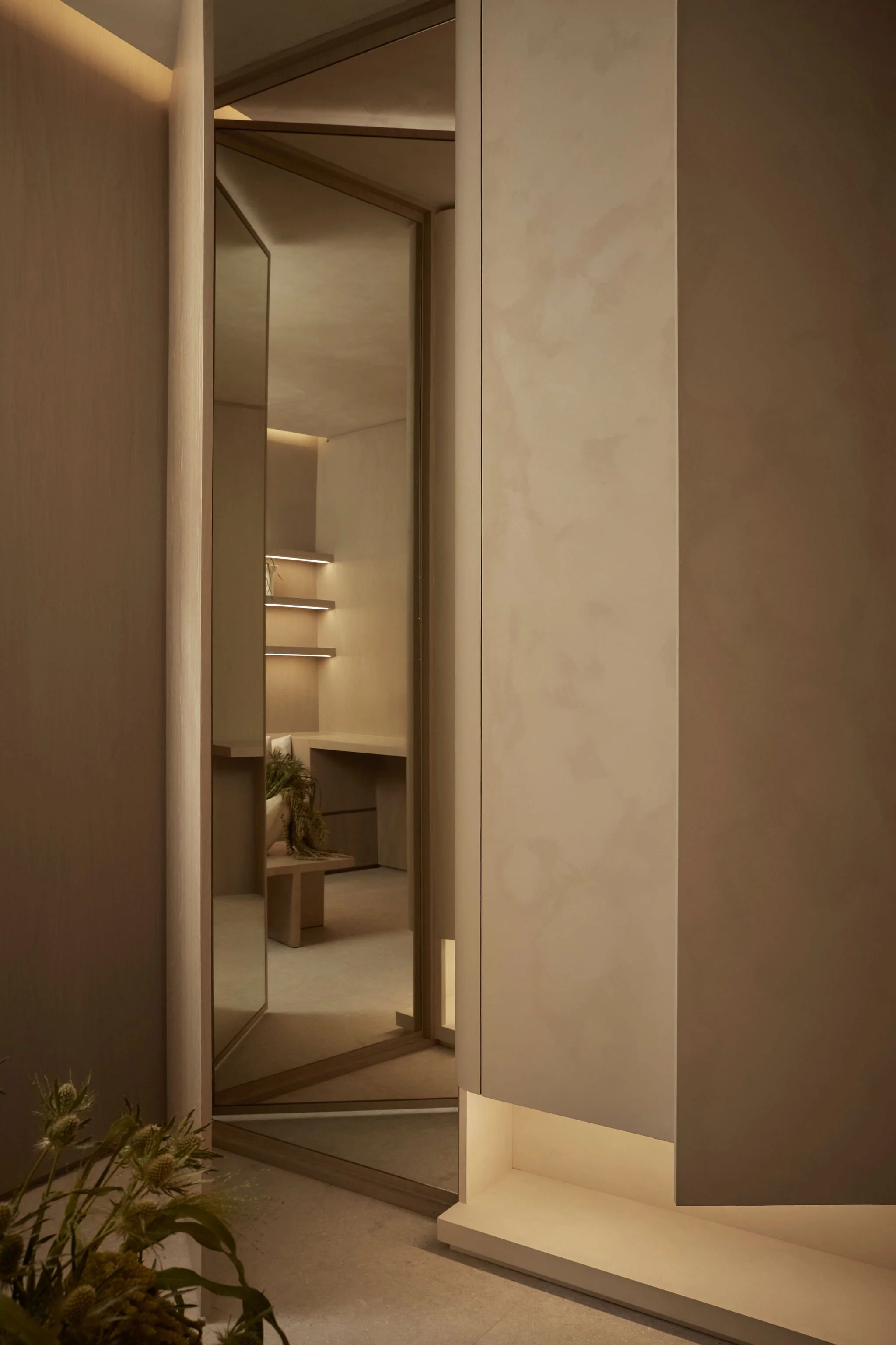 A partially open mirrored door in a modern wellness spa reflecting a warm, minimalist room with illuminated floating shelves, wood paneling, and a built-in vanity desk