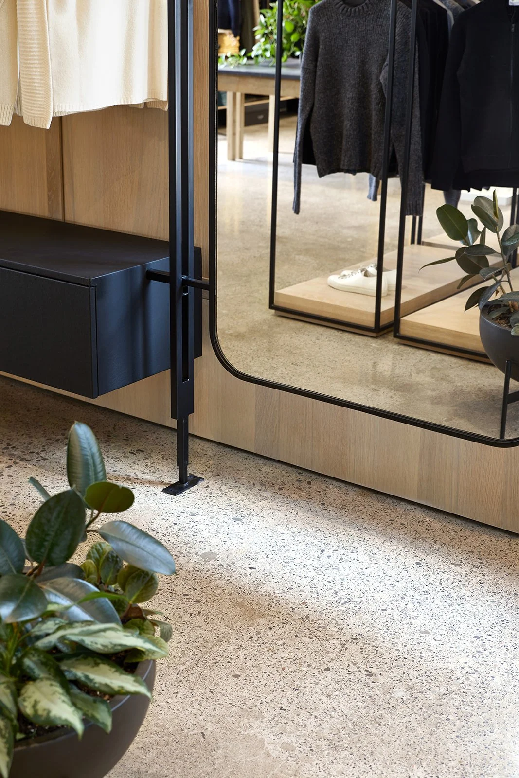 Clothing displayed on racks and a large mirror in a retail store, with plants and a textured concrete floor.