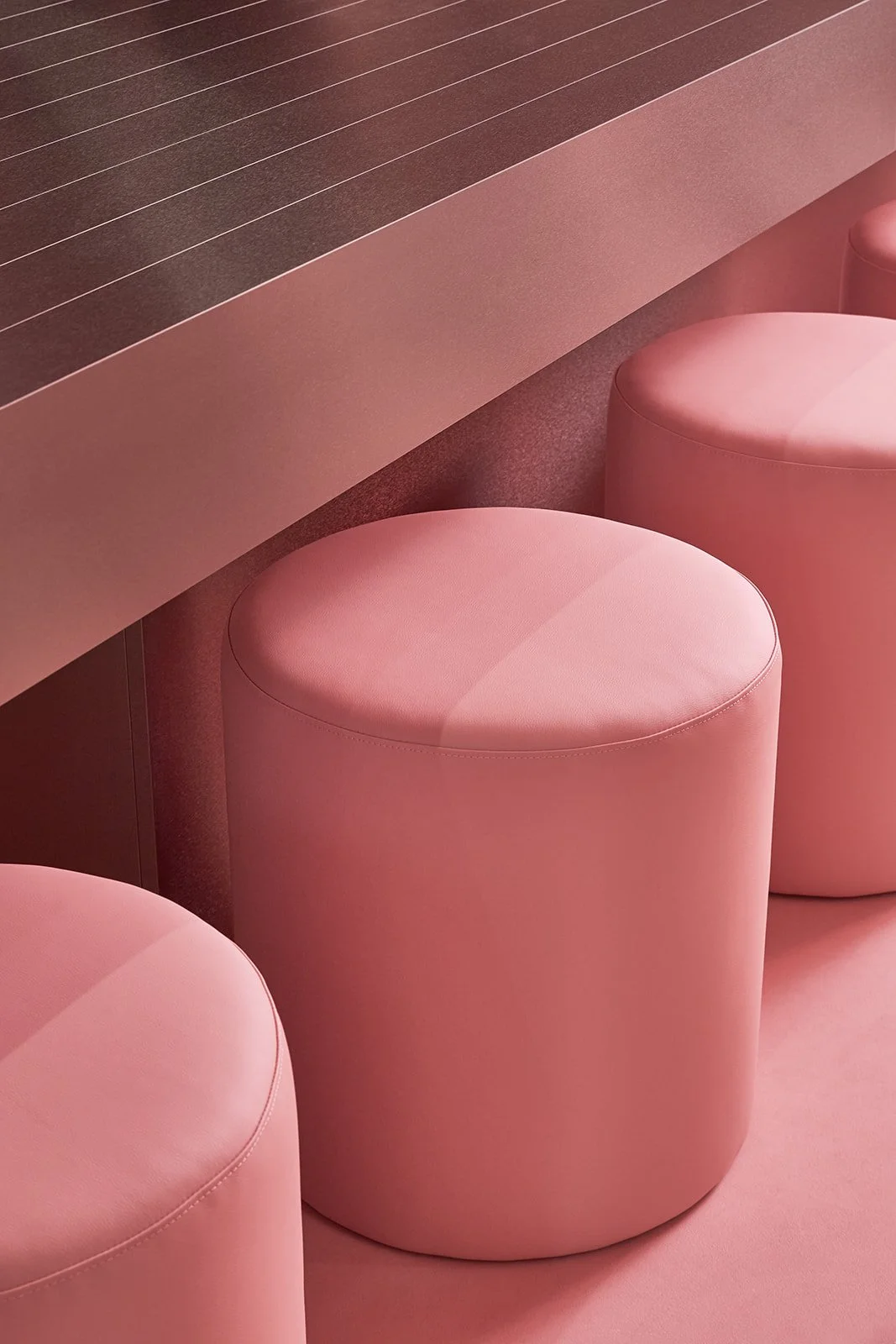 A custom brushed metal cantilevered desk with grid detailing, paired with a plush pink leather stool and a modernist table lamp, set within a bold red and pink anti-clinical wellness space.