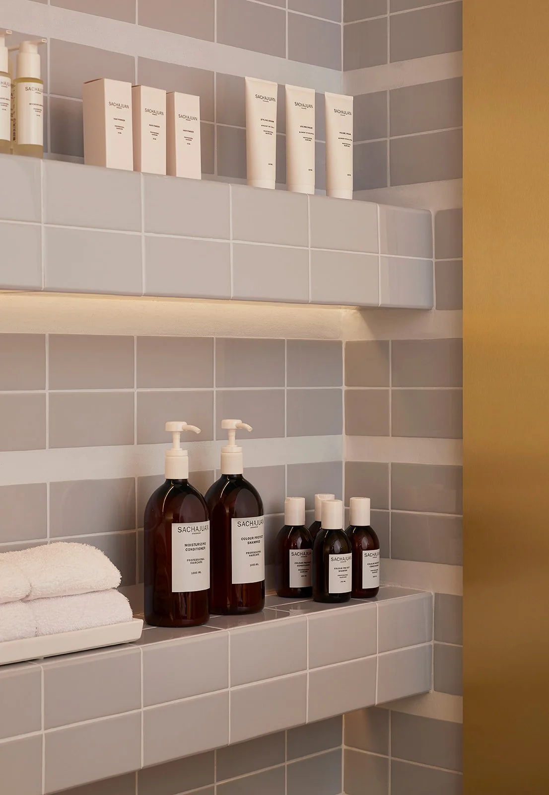 Bathroom shelf holding toiletries, including brown bottles of shampoo and conditioner, white towels, and white boxes of skin care products against tiled wall.