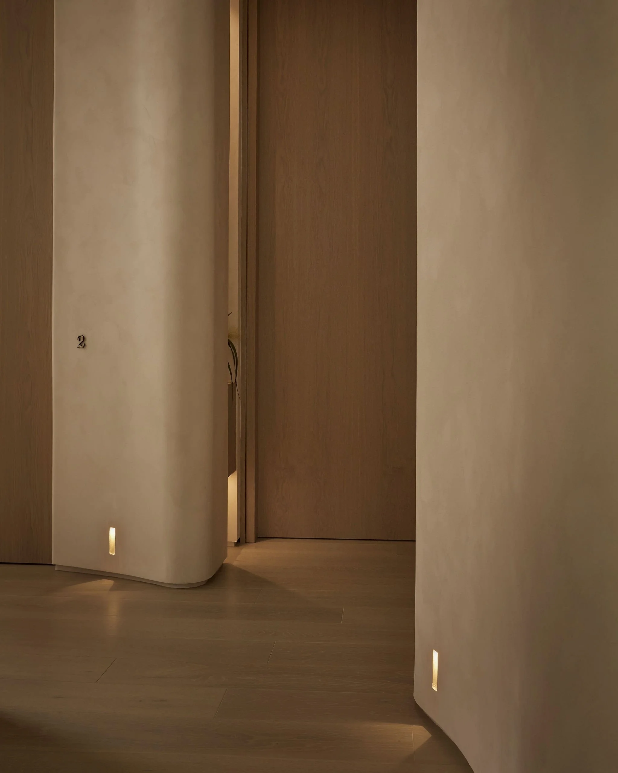 Hallway of a modern wellness clinic featuring thick curved limewash walls, warm wood flooring, a flush wooden door leading to treatment room two, and low-level ambient floor lighting