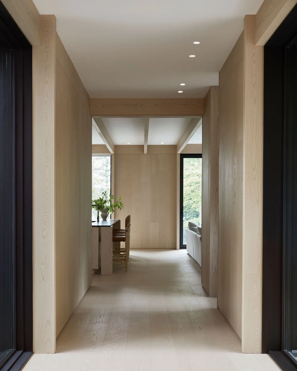 A perspective view down a minimalist hallway in a bespoke recreational retreat, featuring seamless light wood floors, wall paneling, and ceiling beams that perfectly frame a sunlit open-concept kitchen and living space with forest views.